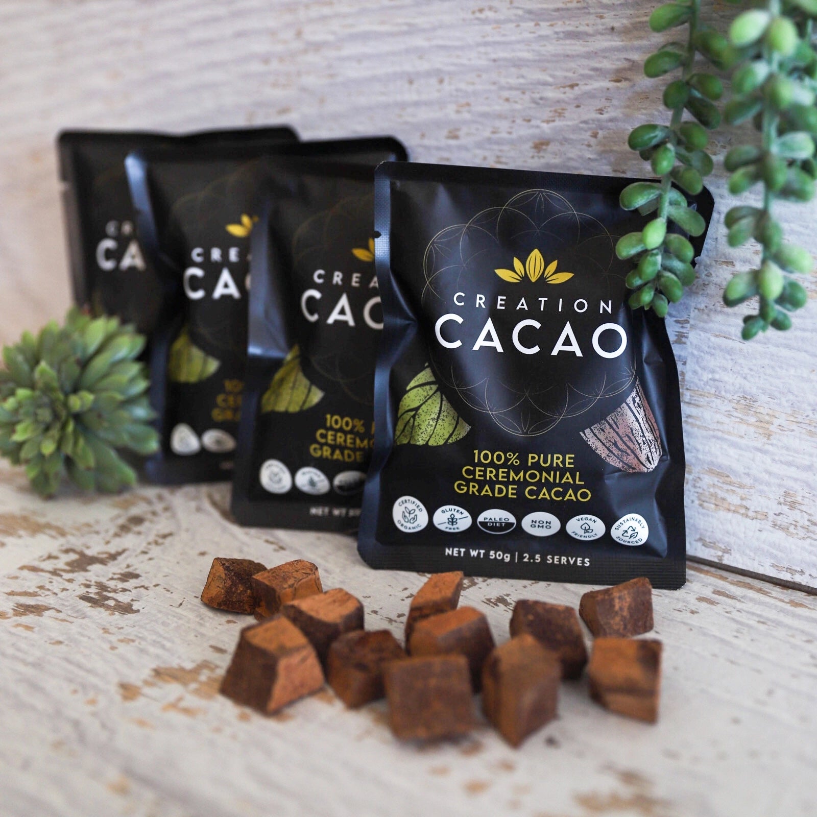 4 packs of the Creation Cacao 50g in front of wooden backdrop and chocolate pieces in front of the pack